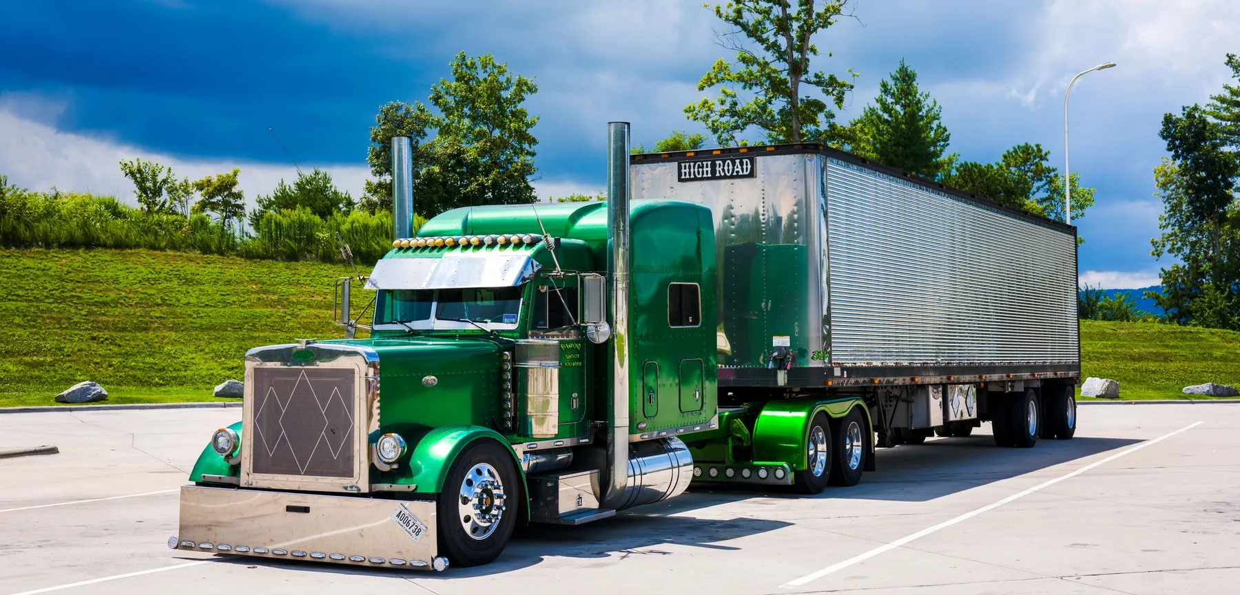 Green conventional semi-truck with a silver dry van trailer parked beside leafy trees.